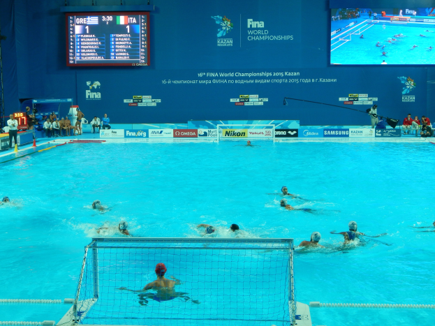 Wasserballspieler bei den 2016 FINA Weltmeisterschaften in Kazan im Pool gegeneinander antretend, mit Zuschauern auf der linken Seite und einem Anzeigebrett mit Event-Logo im Hintergrund.