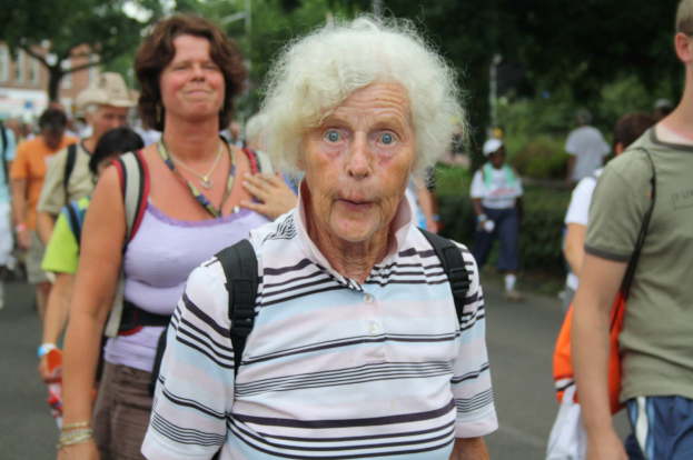 Eine ältere Frau mit öffentlicher Miene geht die Straße entlang, umgeben von Menschen, alle mit Taschen, mit Bäumen, Gebäuden und einem klaren Himmel im Hintergrund.