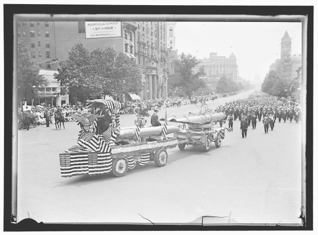 Schwarzes und weißes Foto eines Umzugs mit einem Feuerwehrauto in der Mitte, umgeben von Menschen, einige halten Fahnen, mit Bäumen, Gebäuden mit Fenstern und einer Tafel mit Text im Hintergrund.