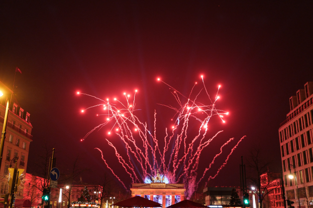 Eine belebte Stadtstraße an einem Silvesterabend in Berlin mit Gebäuden, Bäumen, Laternenmasten, Verkehrszeichen, Zelten, Menschen und einem prächtigen Feuerwerk am Himmel.