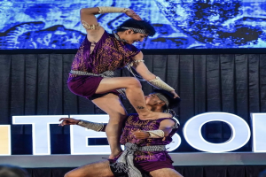 Zwei Performer in violetten Kleidern führen einen akrobatischen Hebemanöver auf der Bühne vor einem Publikum aus, mit einem schwarzen Vorhang und einer Bildschirm-Hintergrund.