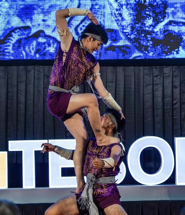 Zwei Performer in violetten Kleidern führen einen akrobatischen Hebemanöver auf der Bühne vor einem Publikum aus, mit einem schwarzen Vorhang und einer Bildschirm-Hintergrund.