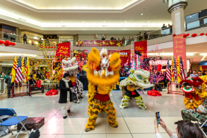 Lebendige Feier des chinesischen Neujahrs in einem Einkaufszentrum mit tanzenden Menschen in Kostümen und anderen, die auf Stühlen sitzen und mobile Telefone halten, umgeben von Fahnen, Bannern, Dekorationen und Deckenleuchten.