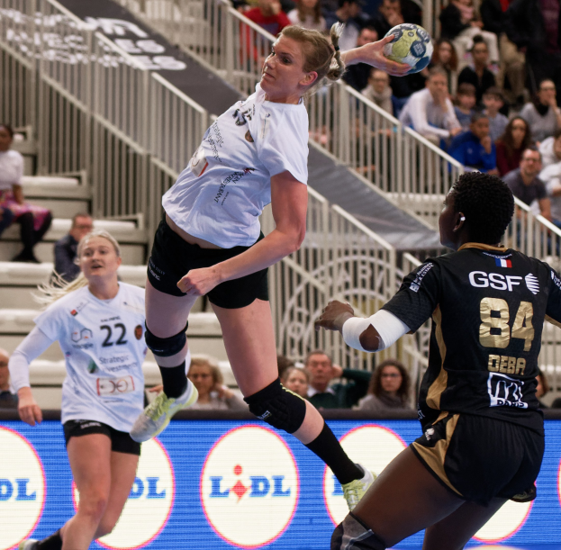Frauen spielen Handball auf einem Feld mit einem Ball in der Luft, Zuschauer im Hintergrund und einer "Handball-Weltmeisterschaft 2019 - Tag 2"-Tafel.