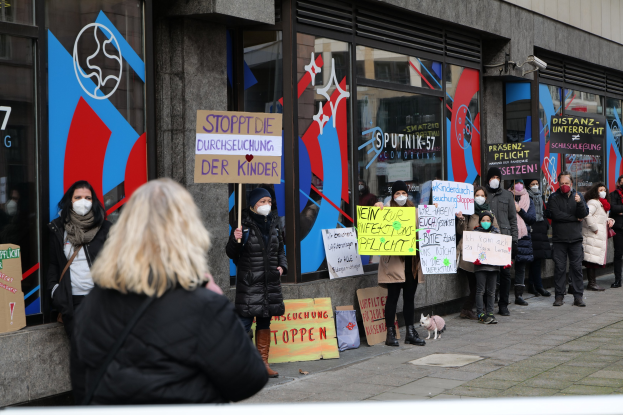 Menschen mit Masken halten Schilder und Plakate und protestieren vor einem Gebäude mit Glastüren, mit Kameras und einem Hund.
