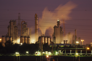 Ölraffinerie bei Nacht mit Rauchentwicklung, beleuchtet von umliegenden Lichtern, mit sichtbarem Himmelhintergrund und einem Wasserzeichen.