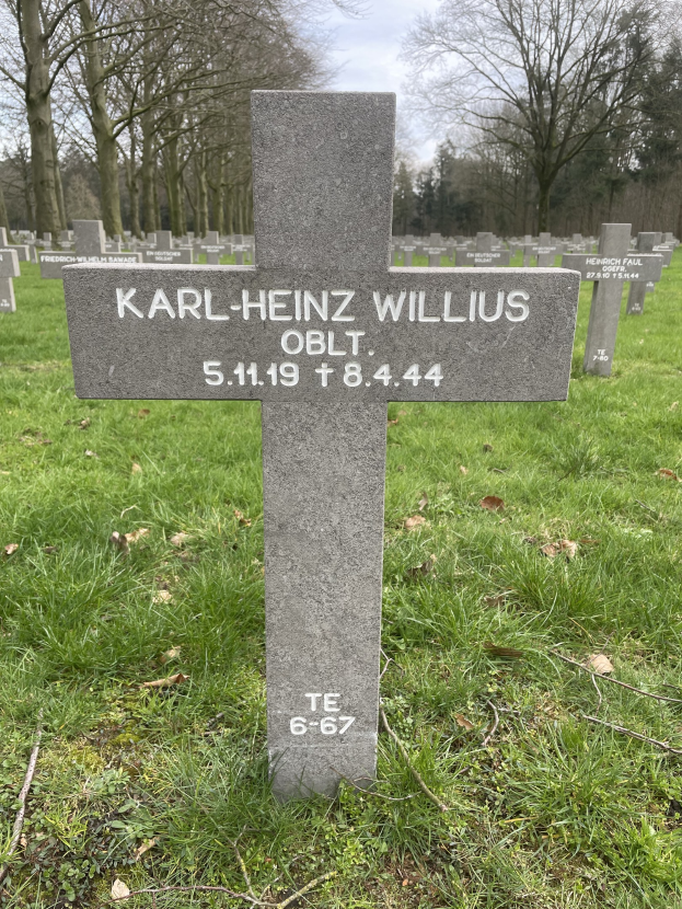 Grab von Karl-Heinz Willis auf einem deutschen Friedhof, umgeben von Gras und trockenen Blättern, mit Bäumen und einem klaren blauen Himmel im Hintergrund.