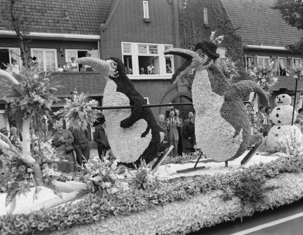 Schwarzes und weißes Foto eines Paradewagens, der mit Blumen und Pinguinfiguren geschmückt ist, mit Menschen in der Nähe und Gebäuden im Hintergrund.