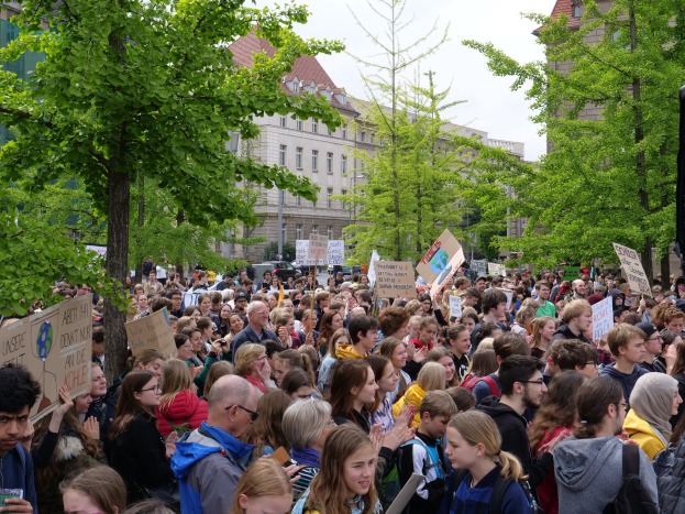 Eine große Menge von Menschen protestiert vor einem Gebäude in Berlin, mit Schildern, mit Bäumen, Fahrzeugen, einem Lautsprecher und Himmel.