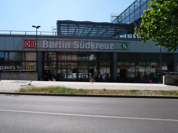 Großes gläsernes Berliner Südkreuz-Bahnhofsgebäude mit Text an seiner Fassade, Straßenlaternen, Lichtern, Fahrzeugen, sitzenden Fußgängern, Fahrrädern, Bäumen und einem klaren blauen Himmel.