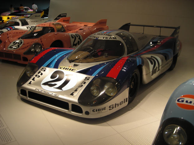 Ein Porsche 917K Rennwagen in einem Museum ausgestellt, umgeben von anderen Sportwagen, mit Menschen in der Nähe und einer Wand im Hintergrund; ein Textschild ist links zu sehen.