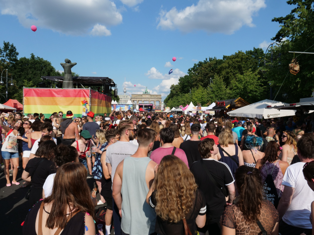 Eine große Menge Menschen geht eine Straße mit Zelten, Bäumen, Pfählen, Lichtern und einer Statue entlang, mit Gebäuden und einem bewölkten Himmel mit Ballons im Hintergrund während des Berliner Christopher Street Day.