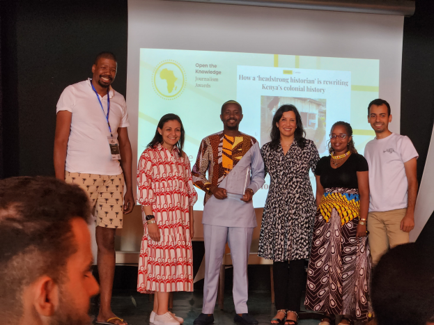 Eine Gruppe von Menschen auf einer B├╝hne, die fr├Âhlich in die Kamera l├Ąchelt und posiert, mit einem Publikum darunter und einem Bildschirm im Hintergrund, der 'Open the Knowledge Journalism Awards 2019' anzeigt.