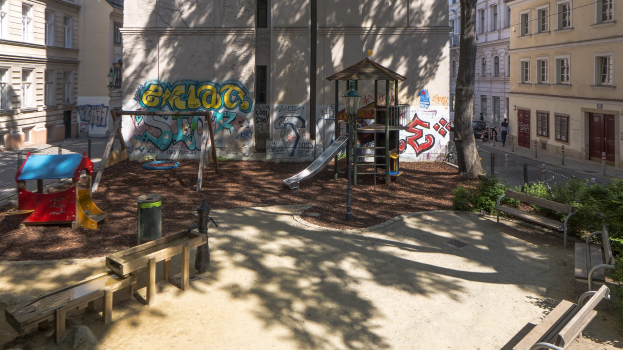 Ein Spielplatz mit Graffiti, umgeben von Gebäuden, Bäumen, Bänken, Pfosten und einem Mülleimer, mit Menschen und Fahrzeugen auf der Straße und dem Himmel im Hintergrund.