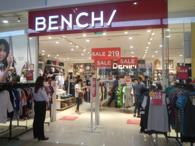 Ein Bekleidungsgeschäft namens "Bench" in einem Einkaufszentrum, das Kleidung an Bügeln zeigt, mit Informationsschildern, Deckenleuchten und einem Bildschirm auf der linken Seite.