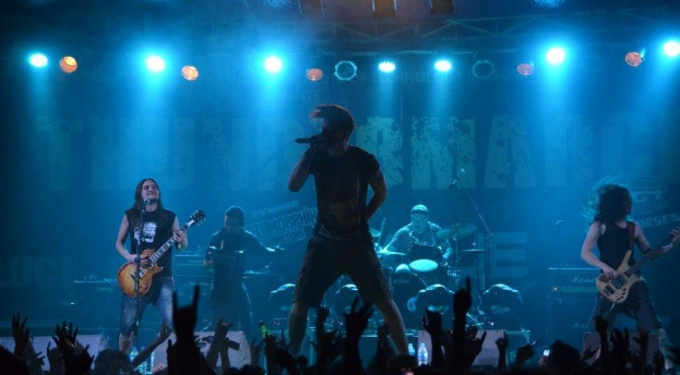 Eine Gruppe von Musikern, die auf einer hell erleuchteten Bühne in der O2 Arena in London spielen, darunter einige mit Gitarren, einer mit Mikrofon und ein Schlagzeuger im Hintergrund, während die Hände des Publikums unten zu sehen sind.