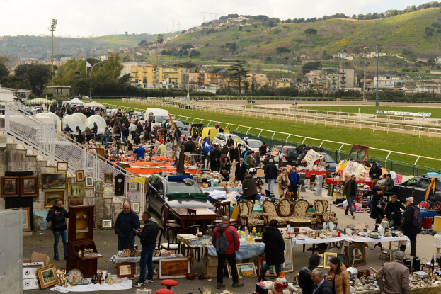 Eine große Gruppe von Menschen auf einem Flohmarkt mit Tischen, auf denen Gegenstände wie Foto Rahmen und Stühle ausgelegt sind, umgeben von geparkten Fahrzeugen, Geländern, Treppen, Bäumen, Gebäuden, Laternenmasten, Hügeln und einem bewölkten Himmel.
