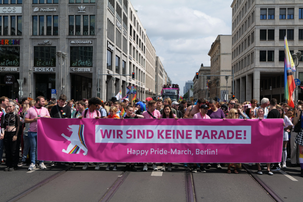 Eine Gruppe von Menschen marschiert auf einer Straße in Berlin, Deutschland, mit einer pinken 'Happy Pride March'-Fahne, während Gebäude, Laternenpfähle und Verkehrszeichen die Straße säumen und der Himmel bewölkt ist.