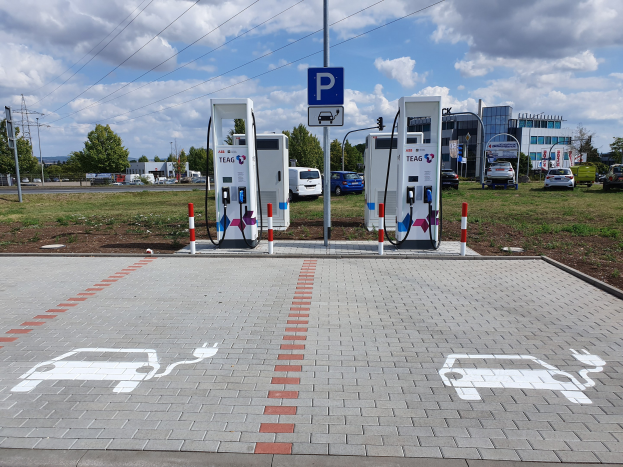 Ladestation für Elektroautos auf einem Parkplatz mit Gebäuden, Bäumen, Strommasten und Fahrzeugen auf der Straße unter einem bewölktem Himmel.