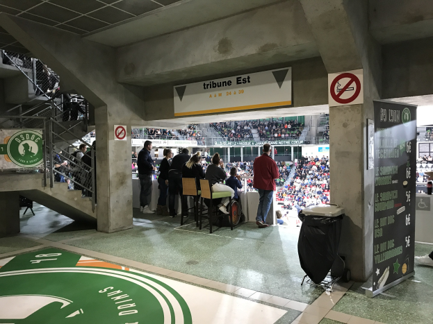 Eine Gruppe von Menschen steht vor einem sitzenden Publikum in einem Stadion während eines Boston Celtics vs New York Jets Ereignisses, mit Schildern, einem Müllcontainer, einer Texttafel, einer Treppe und Deckenlampen.