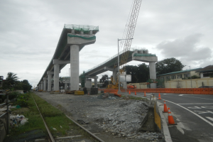 Baustelle mit einer Brücke im Hintergrund, eine Straße mit Verkehrskegeln, verstreute Steine und Gras, eine Eisenbahnschiene auf der linken Seite, Bäume und Gebäude auf beiden Seiten und ein bewölkter Himmel oben.