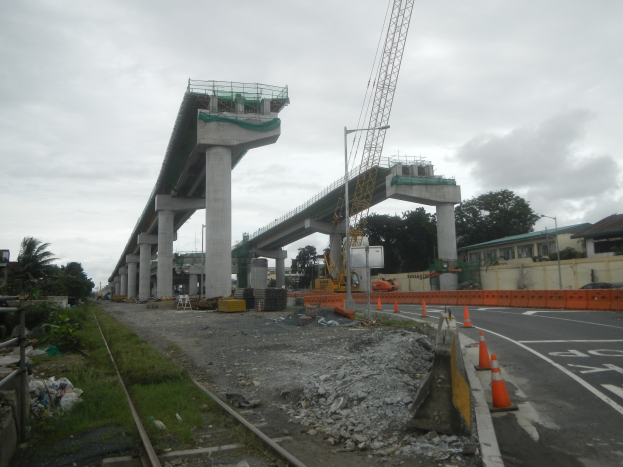 Baustelle mit einer Brücke im Hintergrund, eine Straße mit Verkehrskegeln, verstreute Steine und Gras, eine Eisenbahnschiene auf der linken Seite, Bäume und Gebäude auf beiden Seiten und ein bewölkter Himmel oben.