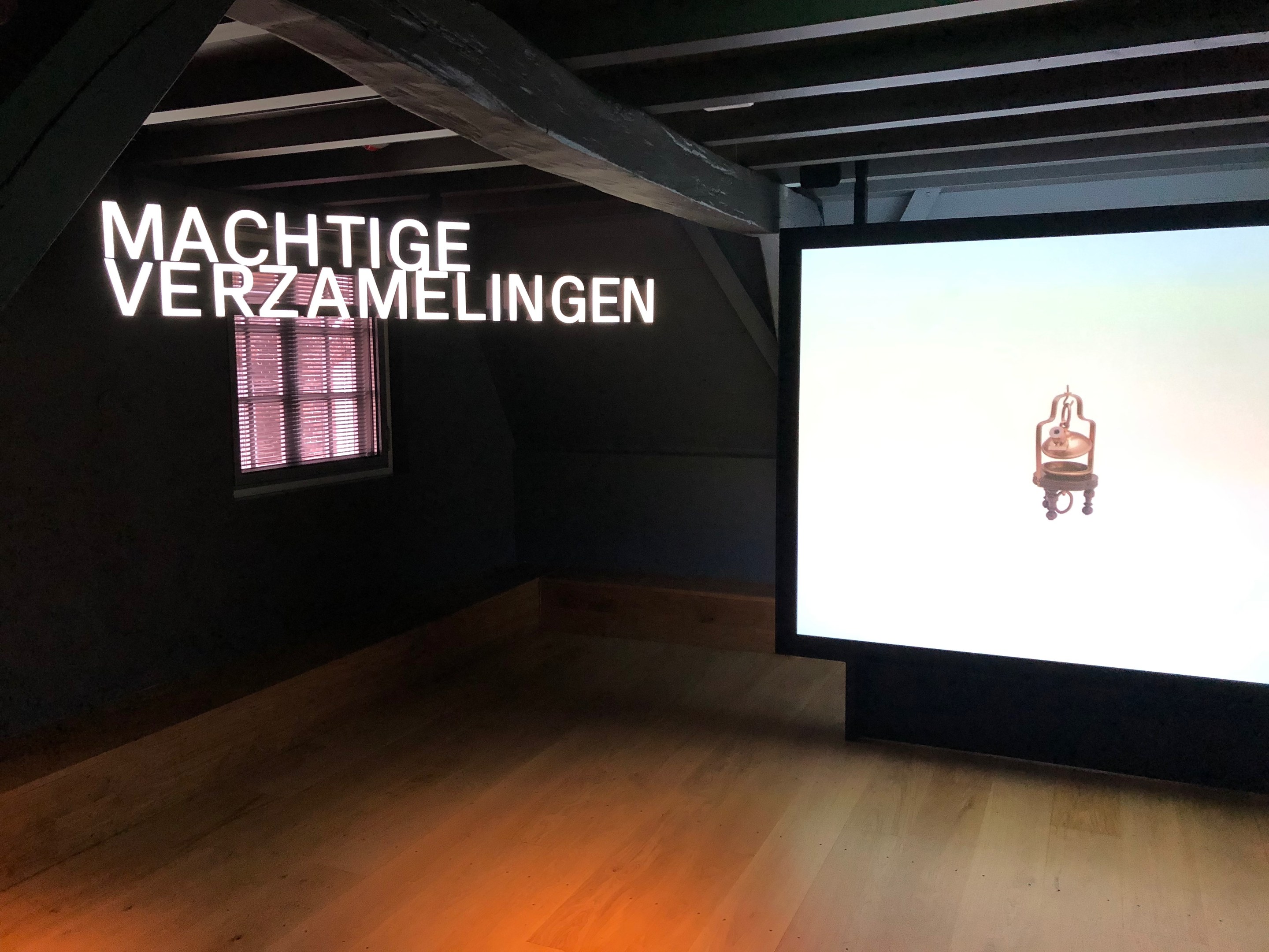 Installationsansicht der Ausstellung "Machtige Verzamelingen" im Museum of Modern Art in Berlin, mit einem großen Bildschirm an der Wand und einem Fenster links.