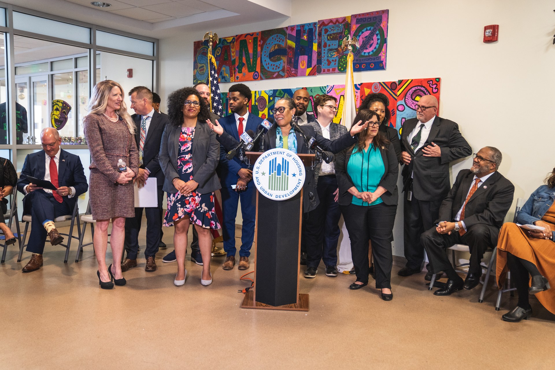 Eine Gruppe von Menschen steht vor einem Podium mit einem Mikrofon und einer Tafel mit der Aufschrift "Sancho Stadtrat joins City Council", während andere auf Stühlen sitzen oder in der Nähe stehen; Flaggen, ein Feuerlöscher und Deckenleuchten sind an der Rückwand zu sehen.