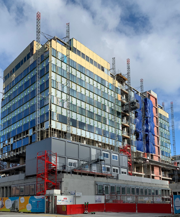 Großes Gebäude im Bau in einer Stadt mit Glasfenstern, Metallrahmen, Treppe, Zaun, Schild, Pfosten, Straßenlaterne, Bäumen und einem bewölkten Himmel.