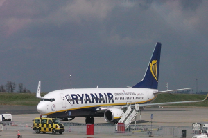 Ein Ryanair-Flugzeug auf dem Rollfeld eines Flughafens, umgeben von verschiedenen Fahrzeugen und Geräten, mit Gras, Bäumen und einem bewölkten Himmel im Hintergrund.