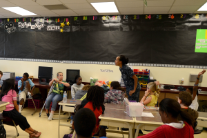 Ein Klassenzimmer mit Kindern an Tischen mit Monitoren, Flaschen, Körben, Papieren und einer Tafel, während ein Lehrer in der Mitte steht, mit Taschen auf dem Boden und Lampen an der Decke.