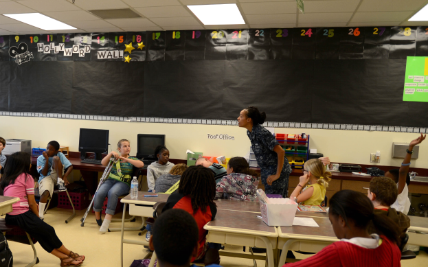 Ein Klassenzimmer mit Kindern an Tischen mit Monitoren, Flaschen, Körben, Papieren und einer Tafel, während ein Lehrer in der Mitte steht, mit Taschen auf dem Boden und Lampen an der Decke.