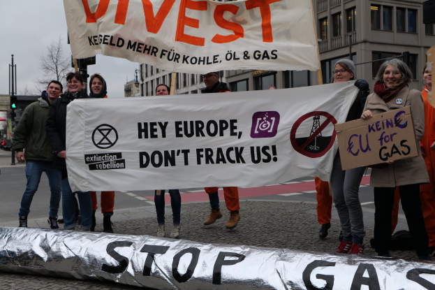 Eine Gruppe von Menschen auf der Straße mit einem Banner "Stoppt Gas" als Protest gegen die Europäische Union, im Hintergrund Gebäude, Bäume, Pfosten, Verkehrszeichen und einen klaren blauen Himmel.