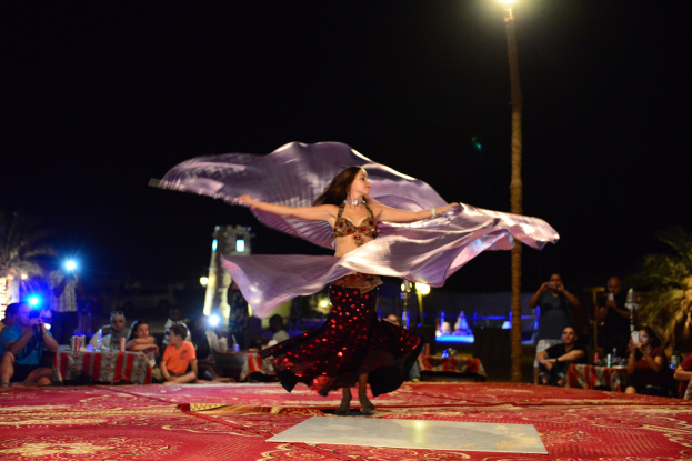 Eine Frau in einem Bauchtanzkostüm tanzt auf einem roten Teppich umgeben von einer Zuschauermenge mit Kameras, Bäumen, Lichtern, einer Stange und Himmel im Hintergrund.