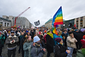 Eine große Gruppe von Menschen bei einer LGBTQ+-Rechtsdemo in Berlin, die Fahnen und Plakate schwingt, mit Gebäuden und einem Kran im Hintergrund unter einem bewölkten Himmel.