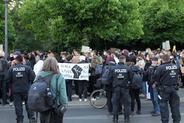 Eine große Gruppe von Menschen steht auf der Straße und hält Schilder und trägt Mützen und Taschen, mit einem Fahrrad im Vordergrund und Bäumen und einem Pfahl im Hintergrund während einer Black Lives Matter Demonstration in Berlin.