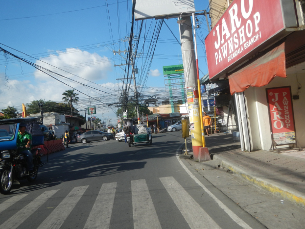 Belebte Stadtstraße mit Fahrzeugen, Fußgängern, Strommasten, Gebäuden, Bäumen und einem bewölkten Himmel, mit einem auf der rechten Seite befindlichen Geschäft mit der Aufschrift "Jaro Pfandleihhaus".