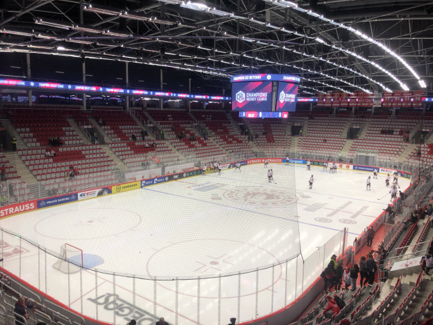 Hockey-Spiel in einer großen Arena mit Zuschauern, eine Eisfläche umgeben von einem Zaun, ein Display, das "St. Louis Blues vs Detroit Red Wings" anzeigt, Metallrahmen, Deckenbeleuchtung und ein Dach.
