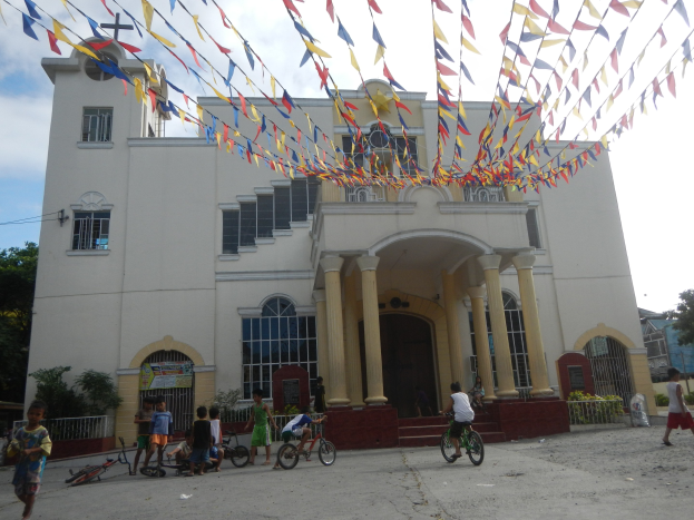 Gruppe von Menschen, die vor einer weißen Kirche mit architectural details, Pflanzen, einem Zaun, einer Fahne, Bäumen und einem bewölkten Himmel Fahrräder fahren, mit bunten Fahnen an einem Seil oben.