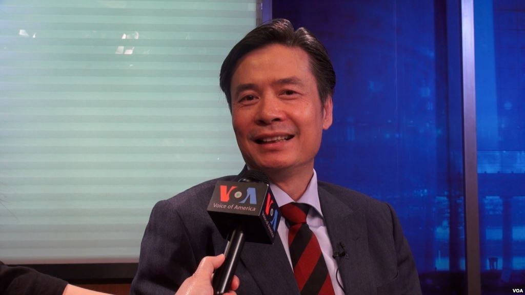 Ein Mann in Anzug und Krawatte hält ein Mikrofon während eines Interviews, wobei die Hand eines Reporters links sichtbar ist, vor einem Fensterhintergrund.