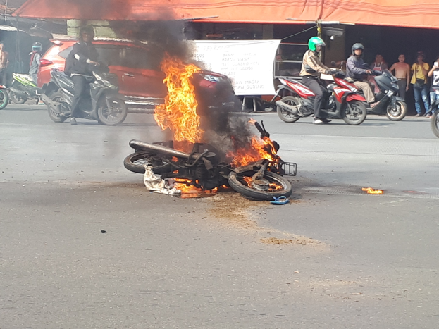 Motorrad in Flammen auf der Straße mit einem Auto und einem Van im Hintergrund, umgeben von Menschen, einige tragen Helme, eine Fahne mit Text und ein Zelt.