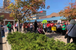 Eine Gruppe von Menschen steht vor einem Gebäude an der University of Michigan, hält Schilder und Kameras hoch, mit Bäumen und Himmel im Hintergrund, protestiert gegen die Schule.