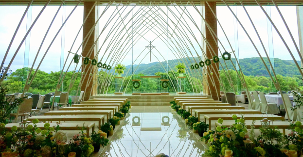 Eine Trauungszeremonie in einem großen Saal mit halbrunder Sitzordnung, geschmückt mit Blumenbouquets und anderen Gegenständen, vor einer Kulisse aus Bäumen, Hügeln und einem klaren blauen Himmel.