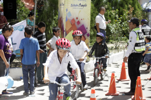 Eine Gruppe von Kindern, die auf Fahrrädern auf einer Straße mit Verkehrskegeln fahren, einige tragen Helme, andere stehen in der Nähe, mit einem Banner, Bäumen und Gebäuden im Hintergrund.