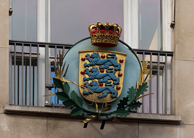 Wappen der britischen Königsfamilie, das auf der Außenseite eines Gebäudes dargestellt wird, begleitet von einem Logo mit Krone und Blättern sowie Fenstern und Metallgittern im Hintergrund.