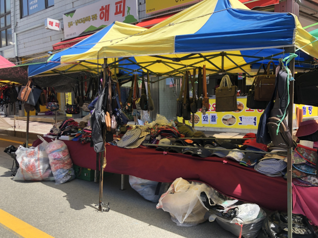 Ein Markt mit Zelten, die Taschen, Hüte und andere Artikel anbieten, vor Häusern mit Fenstern und Texttafeln.