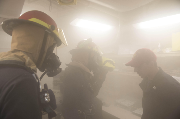 Feuerwehrleute mit Helmen und Rauch vor dem Mund, einer hält eine Kamera, während einer Trainingsrunde mit Hintergrundbeleuchtung und Equipment.