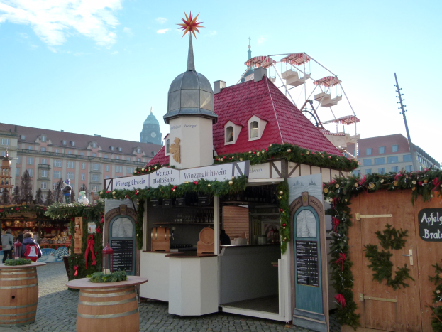 Ein kleines Gebäude mit einem roten Dach und einem Riesenrad davor, umgeben von Menschen und Festschmuck bei einem Outdoor-Event in München, Deutschland.