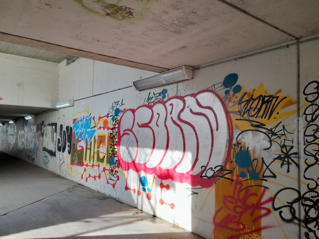 Eine mit Graffiti bedeckte Wand in einer Tiefgarage, beleuchtet von Deckenlampen, präsentiert eine Vielfalt farbenfroher Streetart.