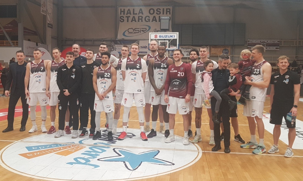 Mannergruppe auf einem Basketballfeld mit Kindern, mit Schildern "Hala Osir Starga", Zuschauern auf Stühlen, einem Basketballkorb, Metallpfosten, Deckenlampen und einer Wand im Hintergrund.